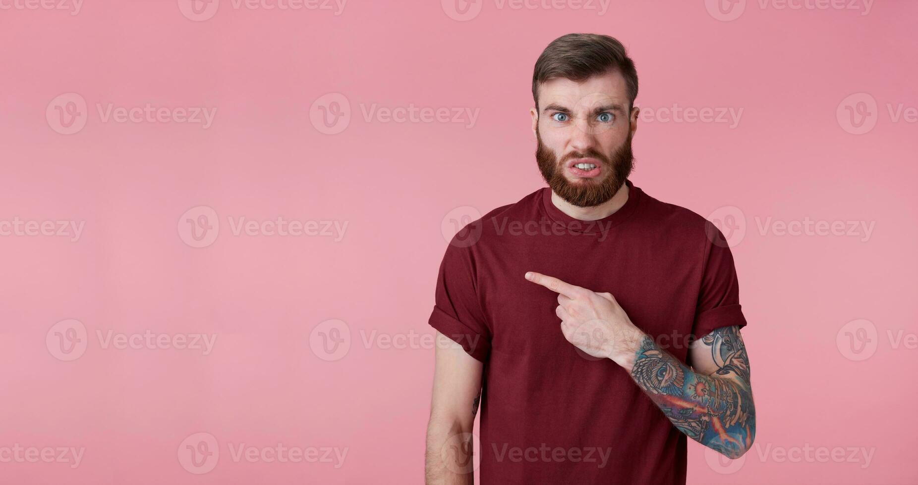Discontented young handsome red bearded man in red shirt, wants to draw your attention to copy space on the left side, frowns face in displeasure, stands over pink background. photo