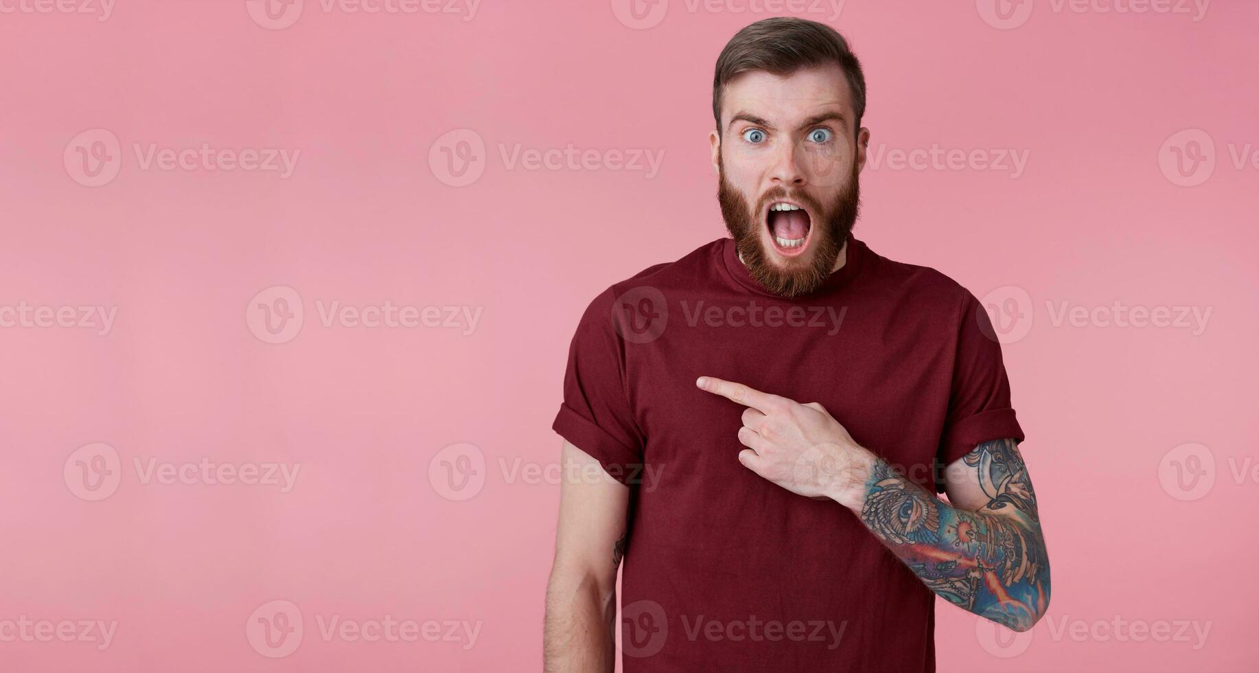 Shocked young handsome red bearded man in red shirt, wants to draw your attention, pointing fingers to the left at the copy space, stands with wide open mouth over pink background. photo