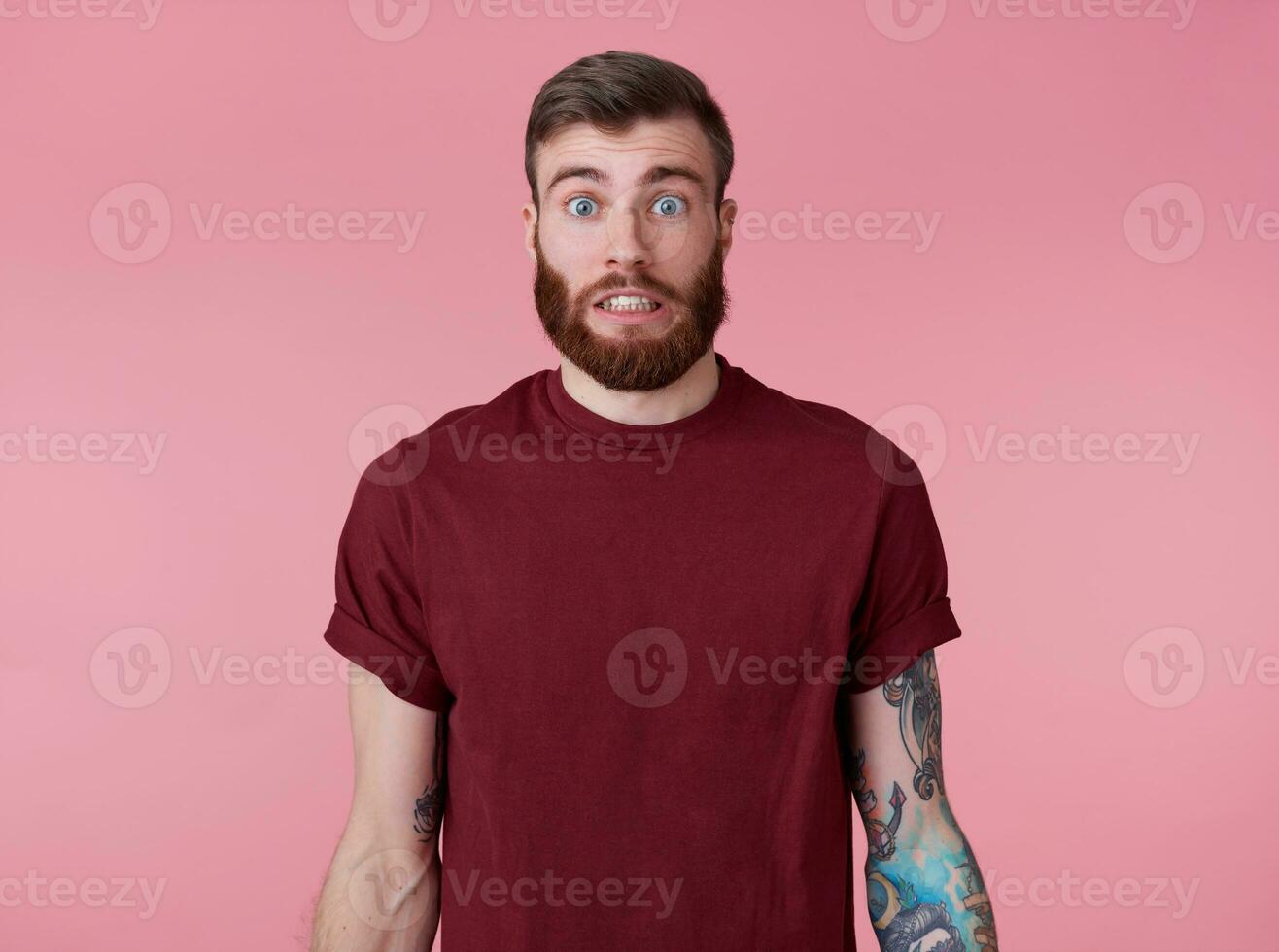 Something wrong Young attractive tattooed red bearded man in blank t-shirt, looks shocked and sad, stands over pink background. photo