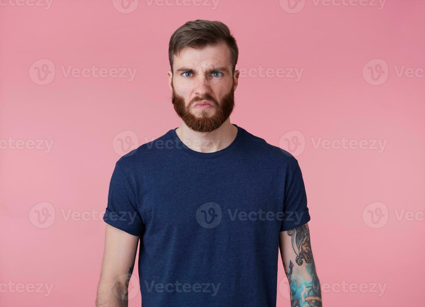 Portrait of young tattooed angry red bearded man in blank t-shirt, frowning and looks at the camera with disgusted, stands over pink background. photo