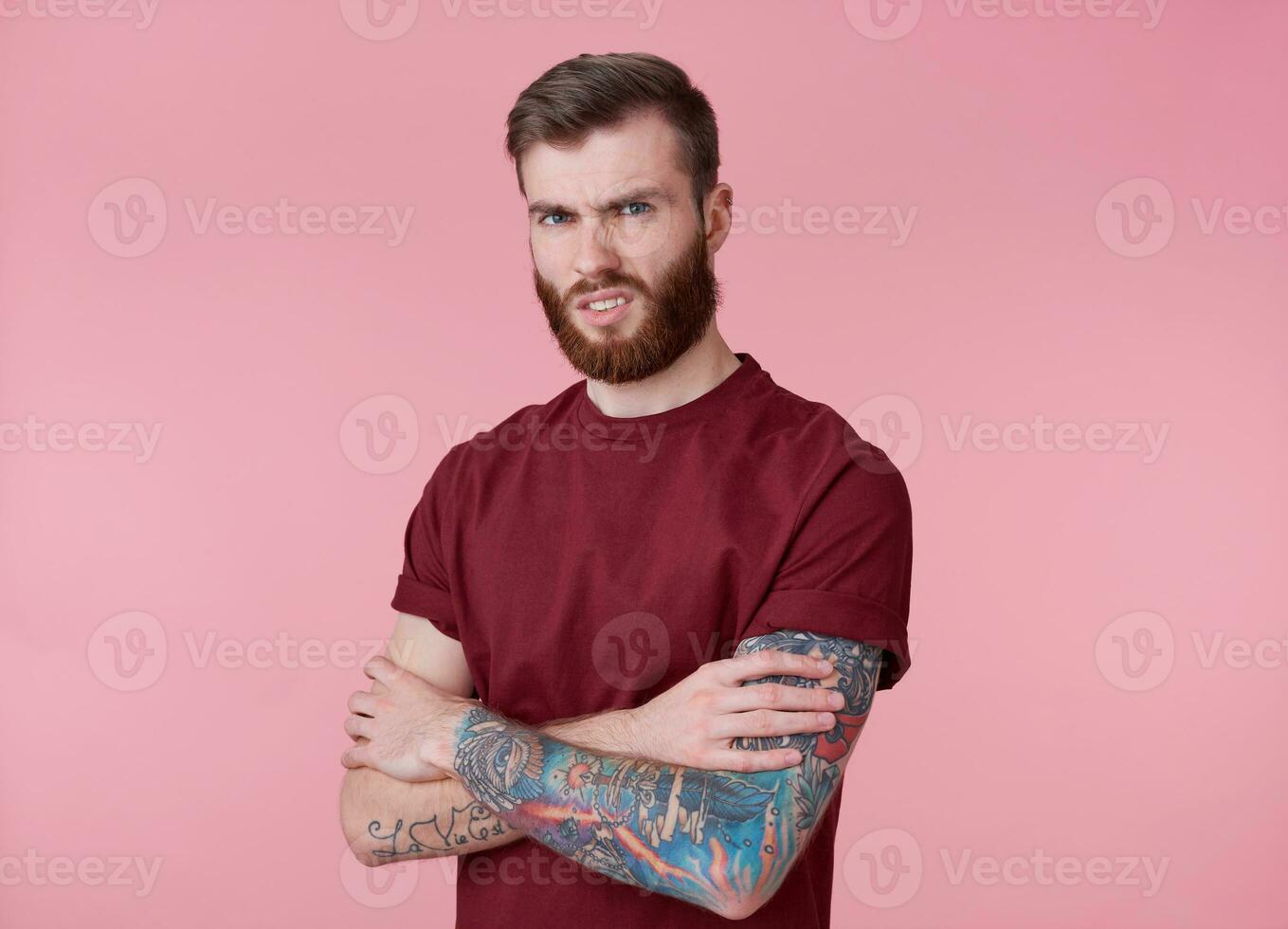 Portrait of young tattooed disgusted red bearded man in blank t-shirt, stands with crossed arms over pink background, frowning and looks at the camera. photo