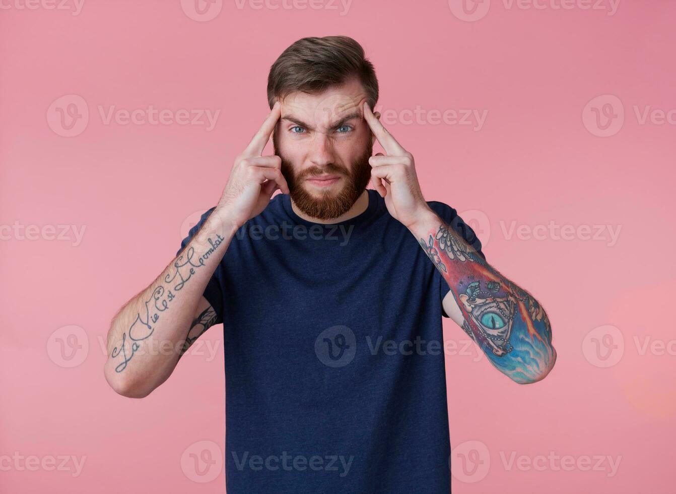 retrato de joven tatuado rojo barbado hombre en blanco camiseta, siente dolor de cabeza y toques templos, soportes terminado rosado antecedentes y ceñudo. foto