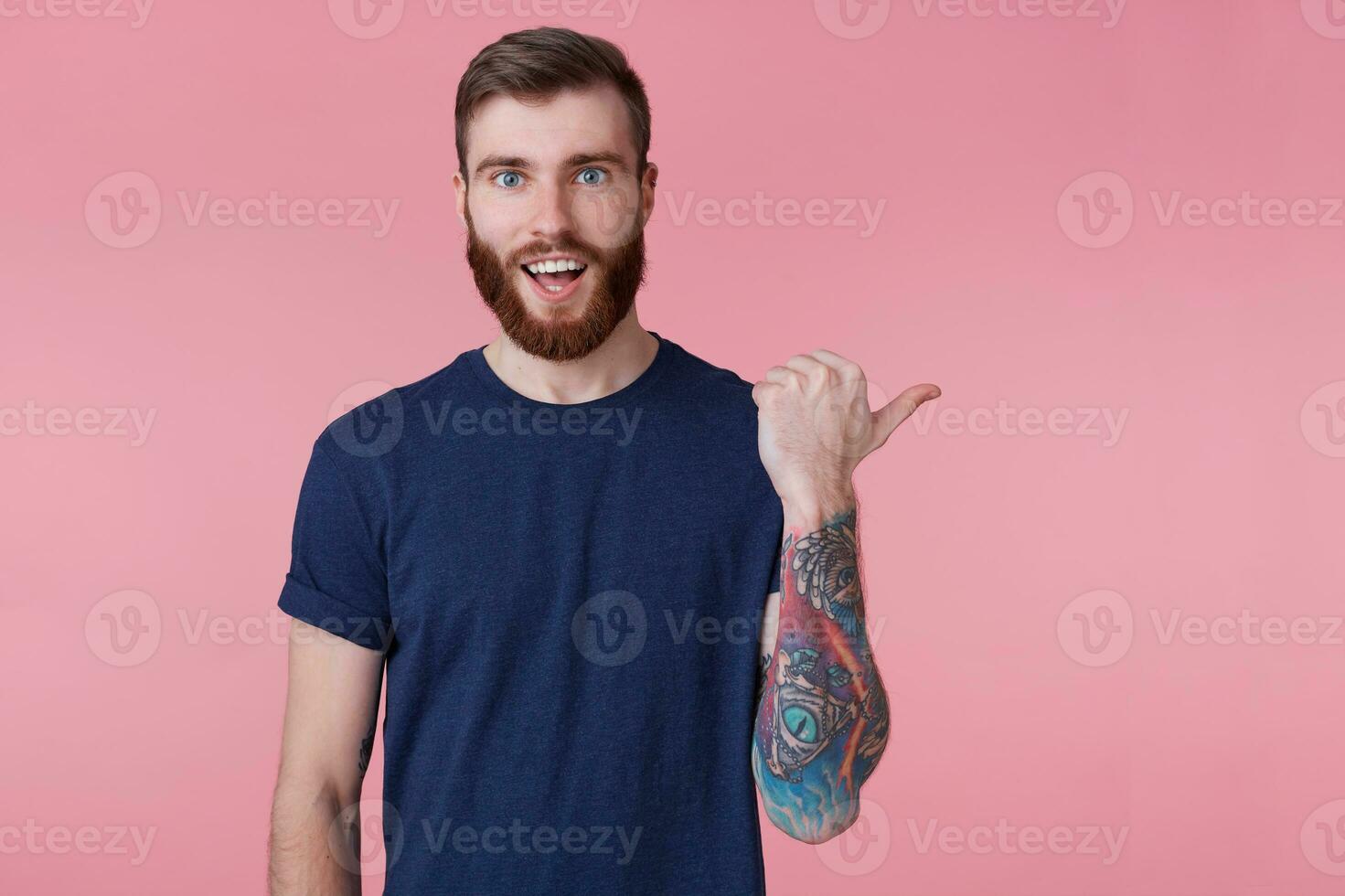 Prttrait of young happy amazed red-bearded young guy ,with wide open mouth in surprise, wearing a blue t-shirt, pointing finger to copy space at the right side isolated over pink background. photo