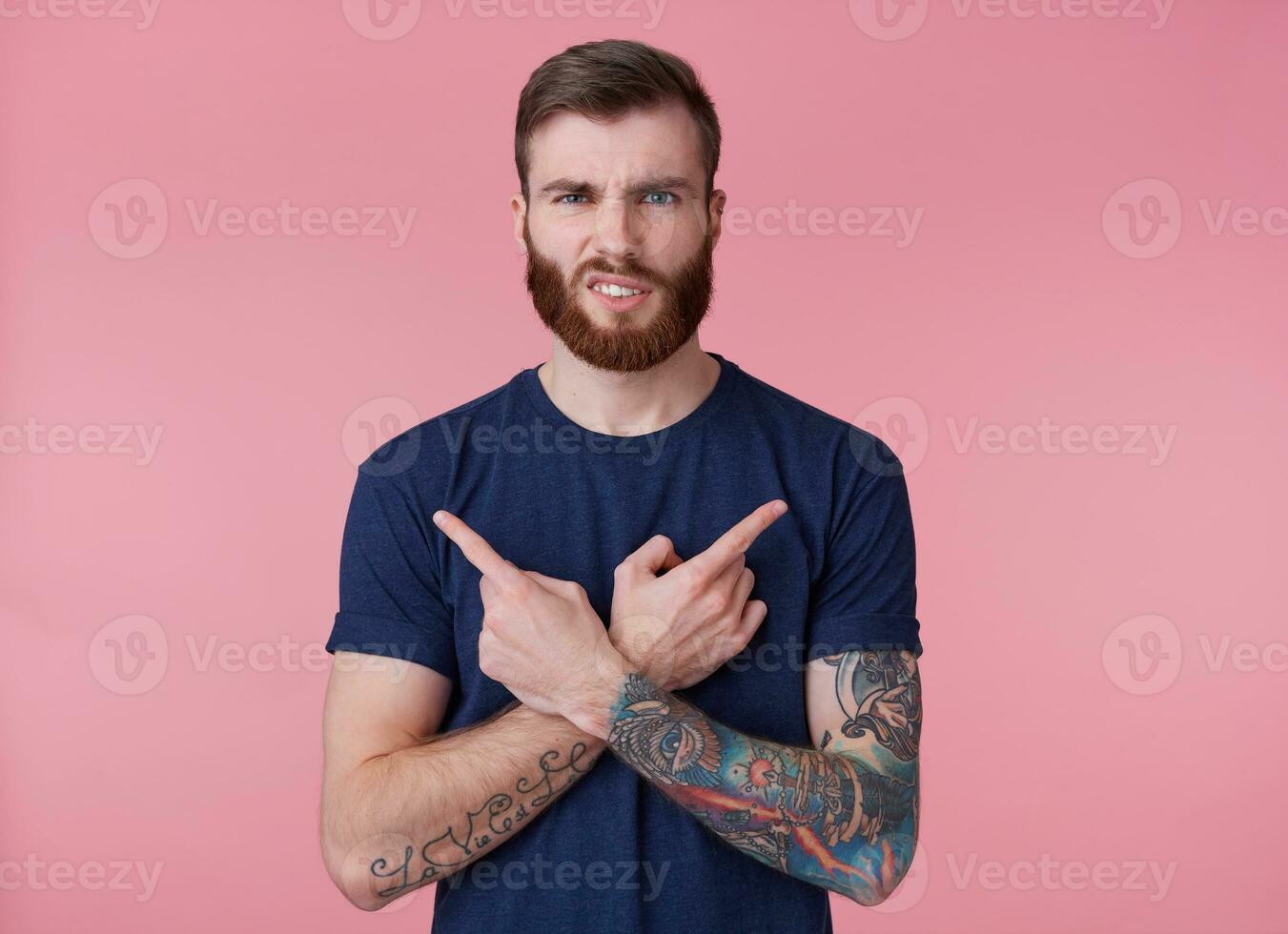 joven de barba roja ceñudo chico , engañado, dedos cruzado, dedos señalando en diferente direcciones, aislado terminado rosado antecedentes. foto