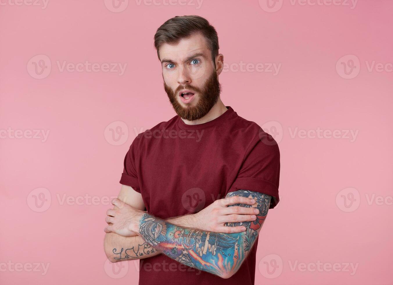 retrato de joven tatuado preguntado rojo barbado hombre en blanco camiseta, soportes con cruzado brazos terminado rosado fondo, mira a el cámara con abierto boca. foto