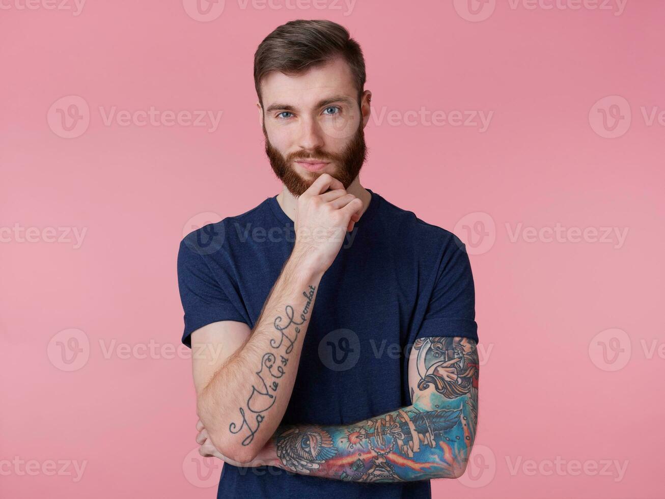 pensativo atractivo de barba roja chico con azul ojos, vistiendo un azul camiseta, sostiene su mano a su barbilla y mira pensativamente dentro el cámara aislado terminado rosado antecedentes. foto