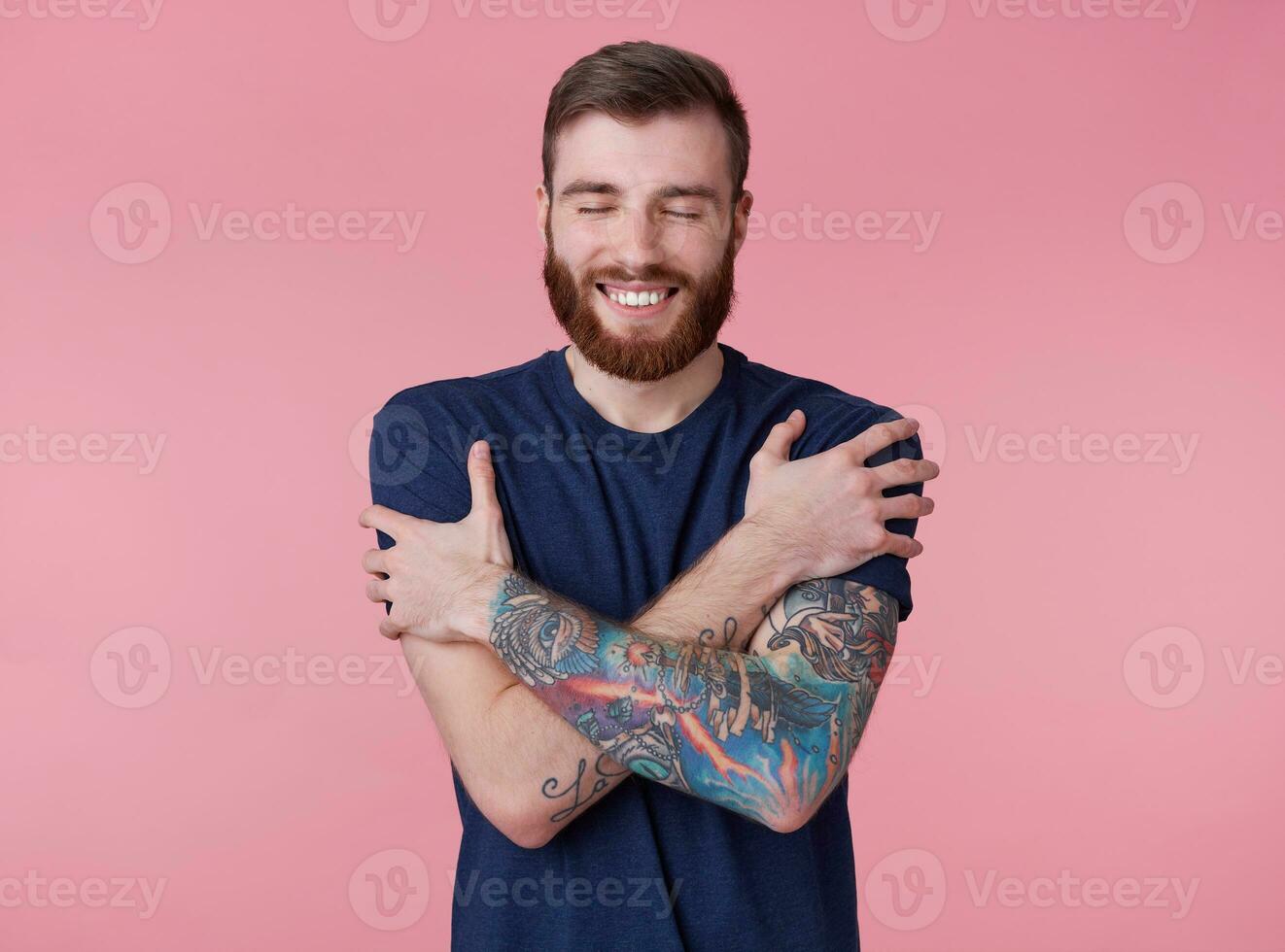 Young happy attractive red-bearded guy with closed eyes, wearing a blue t-shirt, broadly smiling, hugs himself and feels protected isolated over pink background. photo