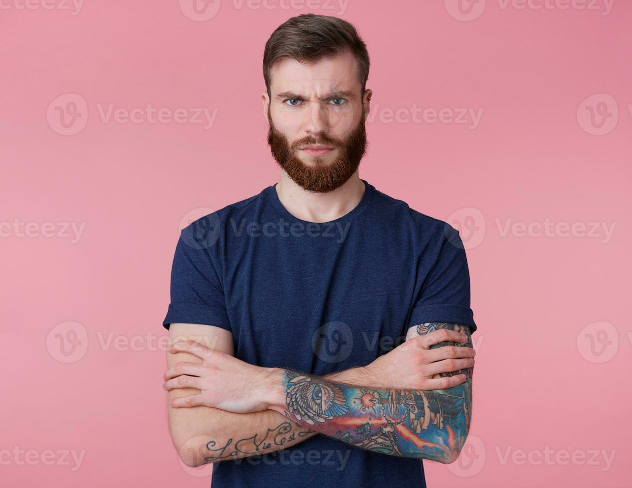 Portrait of severe young red-bearded handsome manful guy with crossed arms, looks outraged, frowning and looking at the camera isolated over pink background. photo