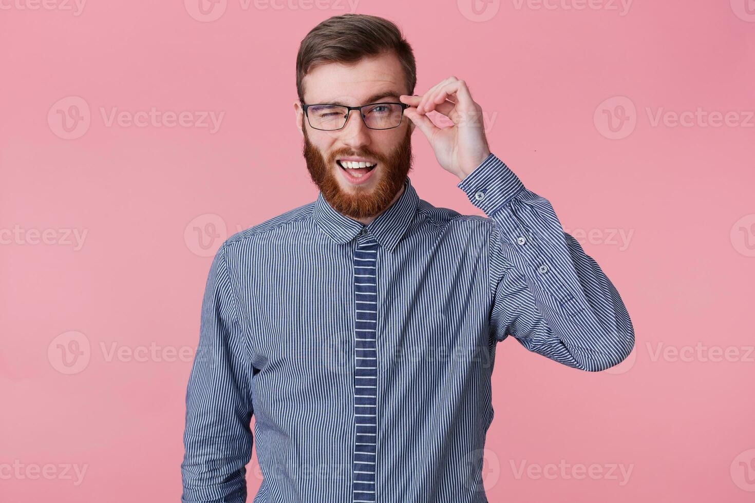 Portrait of young smiling attractive bearded man in a striped shirt, holds glasses and winks isolated over pink background. photo