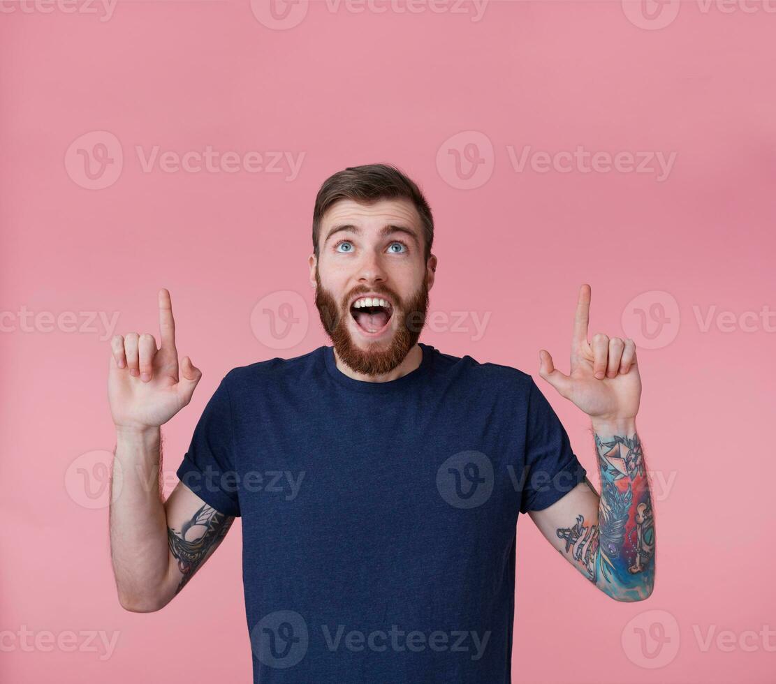 Young happy red-bearded young guy , with wide open mouth in surprise, wants to draw your attention pointing fingers up to copy space isolated over pink background. photo