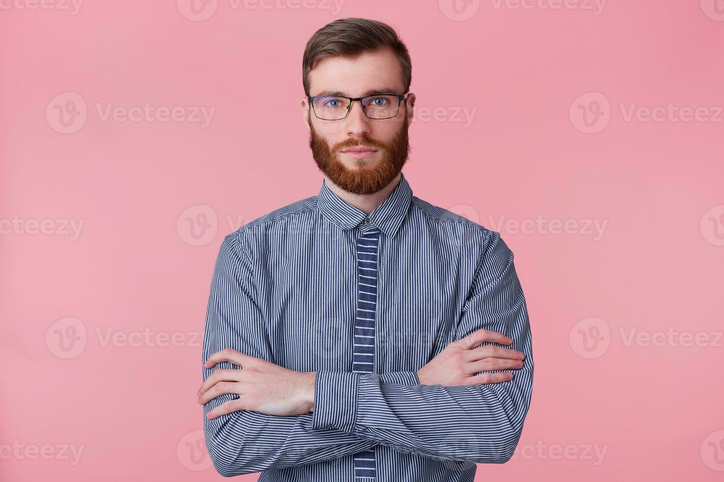 Portrait of a calm bearded young man wears a striped shirt, keeps his arms crossed and looking at the camera isolated on a pink background. photo