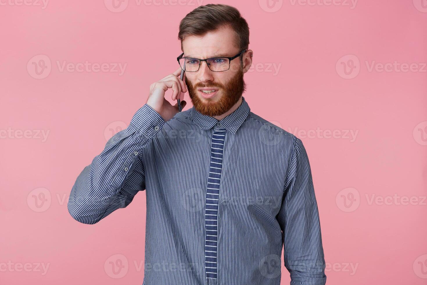 Studio photo of a perplexed young bearded man with glasses dressed in a striped shirt, who is told something unpleasant by phone. Isolated over pink background.