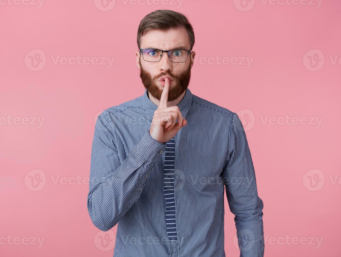 Young handsome red bearded man with glasses and a striped shirt, keeps finger on lips, tells secret information, demonstrates hush gesture isolated over pink background. photo