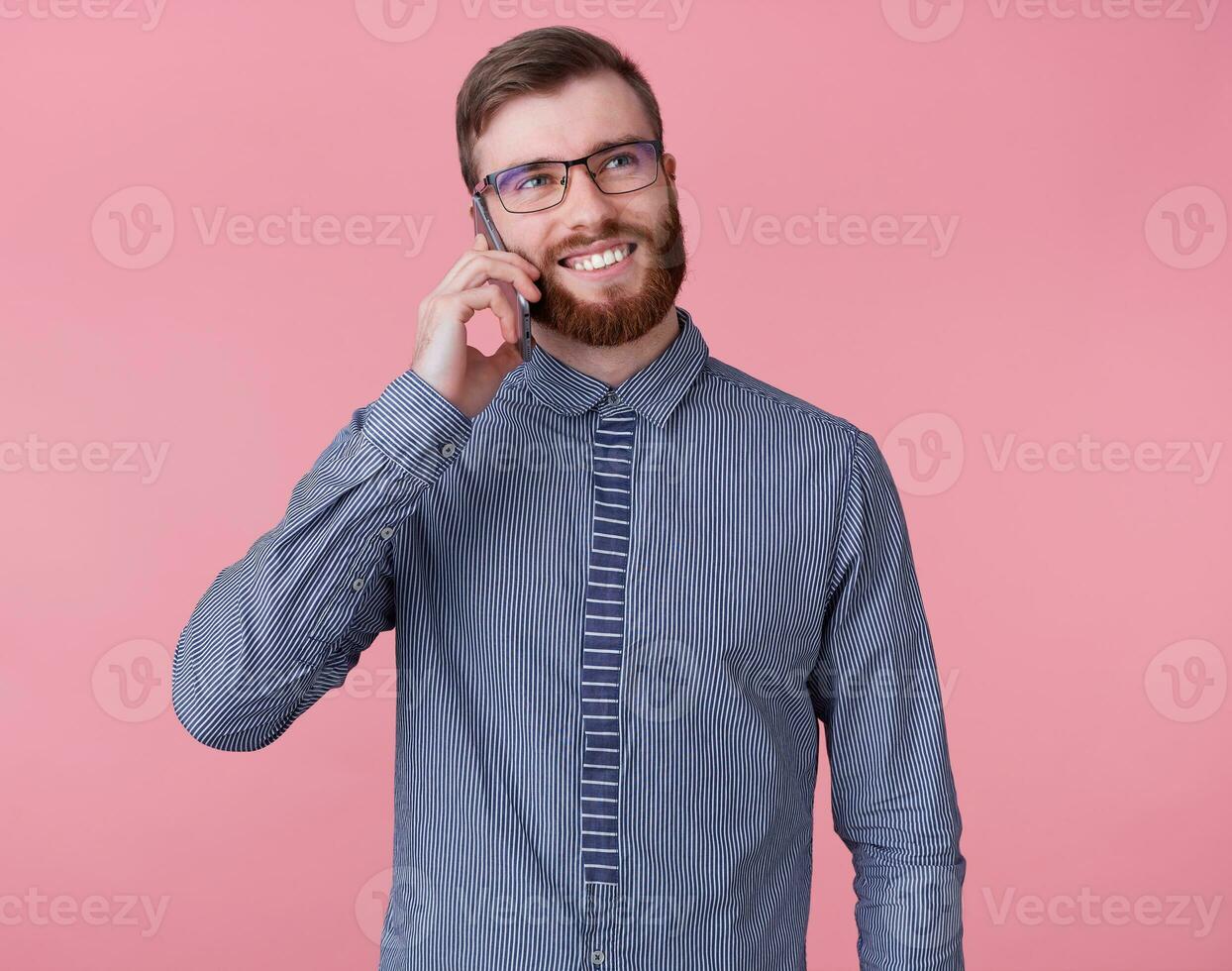 Young attractive red-bearded man smiles broadly and looks up, talking on the phone with his girlfriend, stands over pink background. photo