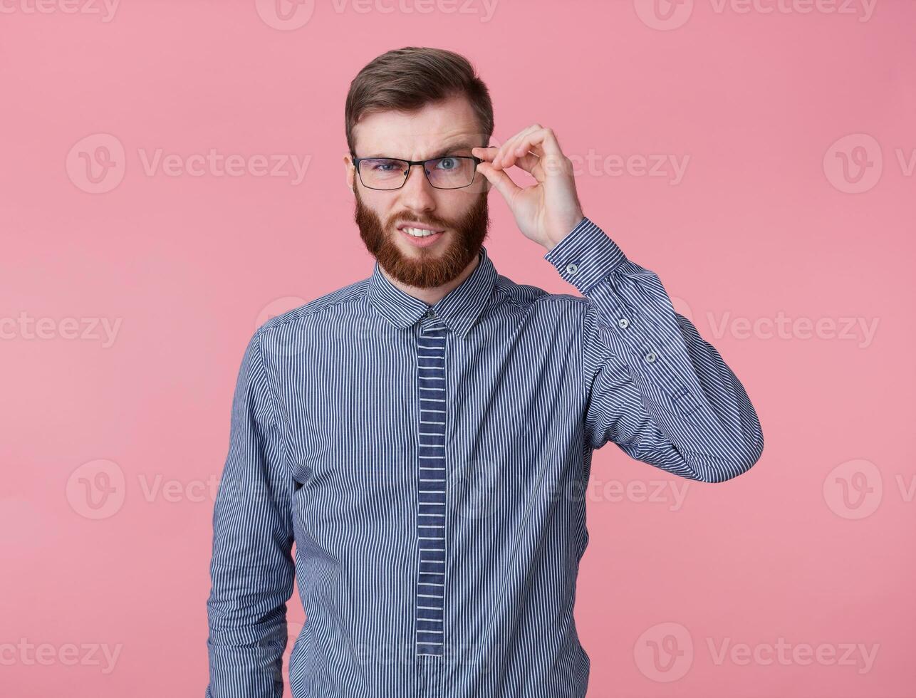 Displeased handsome young handsome red bearded man with glasses and a striped shirt, stands over pink background, raising his eyebrow questioningly, doubts and looks at the camera. photo