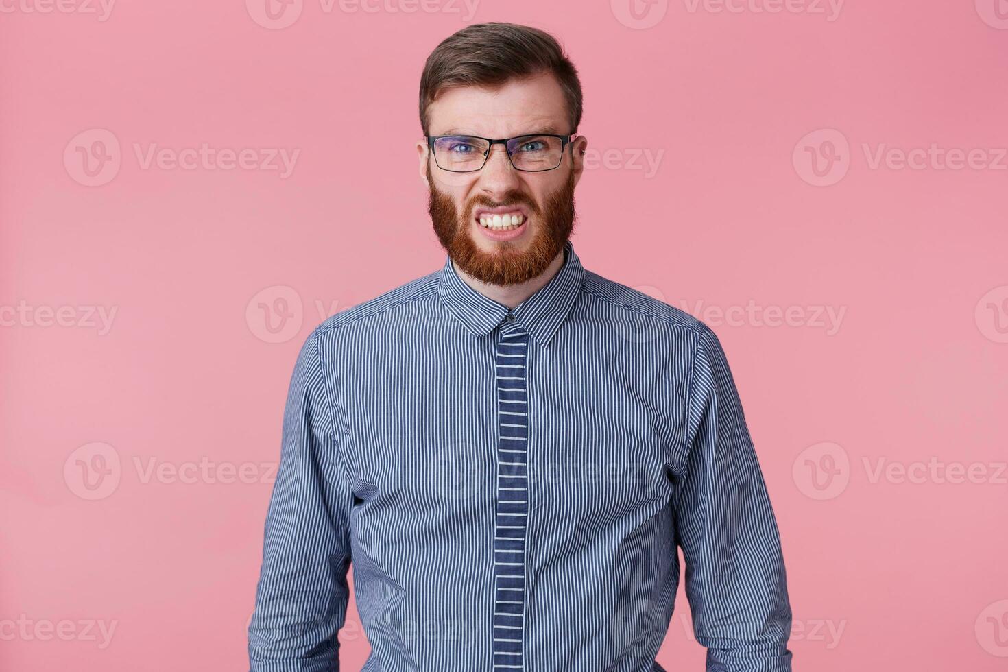 Portrait of young bearded man in a striped shirt with glasses, angry and aggressively bares his teeth isolated over pink background. photo