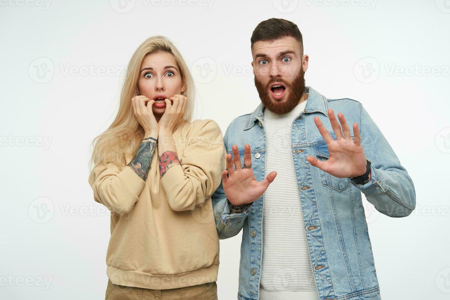 Scared young pretty couple rounding amazedly their blue eyes and raising emotionally hands while looking dazedly at camera, isolated over white background photo