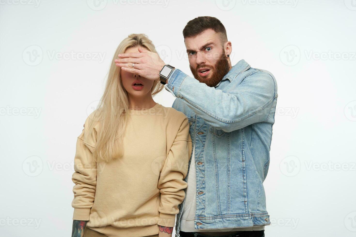 Bewildered young handsome short haired brunette bearded man raising his hand while closing eyes of his blonde surprised girlfriend, isolated over white background photo