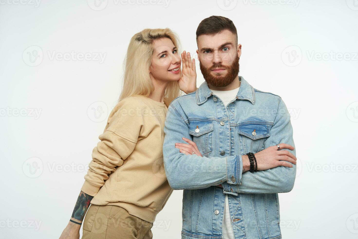 Indoor photo of young severe brown haired bearded man frowning eyebrows while his positive blonde pretty girlfriend telling him something, standing over white background