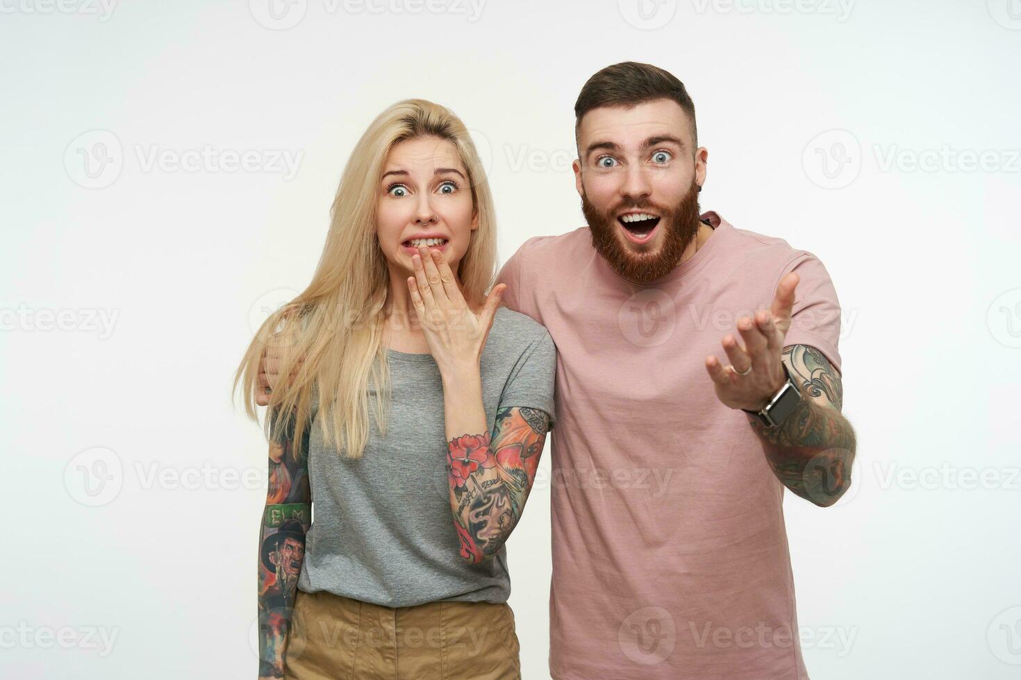 Studio photo of young attractive tattooed couple raising excitedly hands and looking amazedly at camera with wide eyes opened, isolated over white background