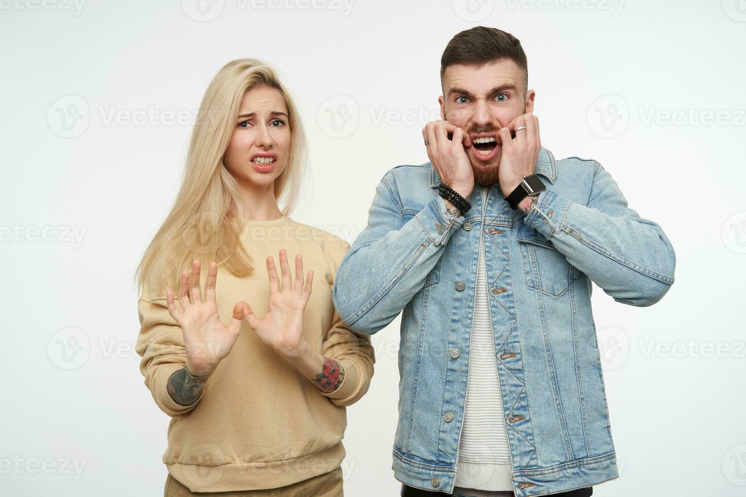 Attractive young long haired blonde female grimacing her face and raising hands in protective gesture while posing with shocked short haired brunette guy over white background photo