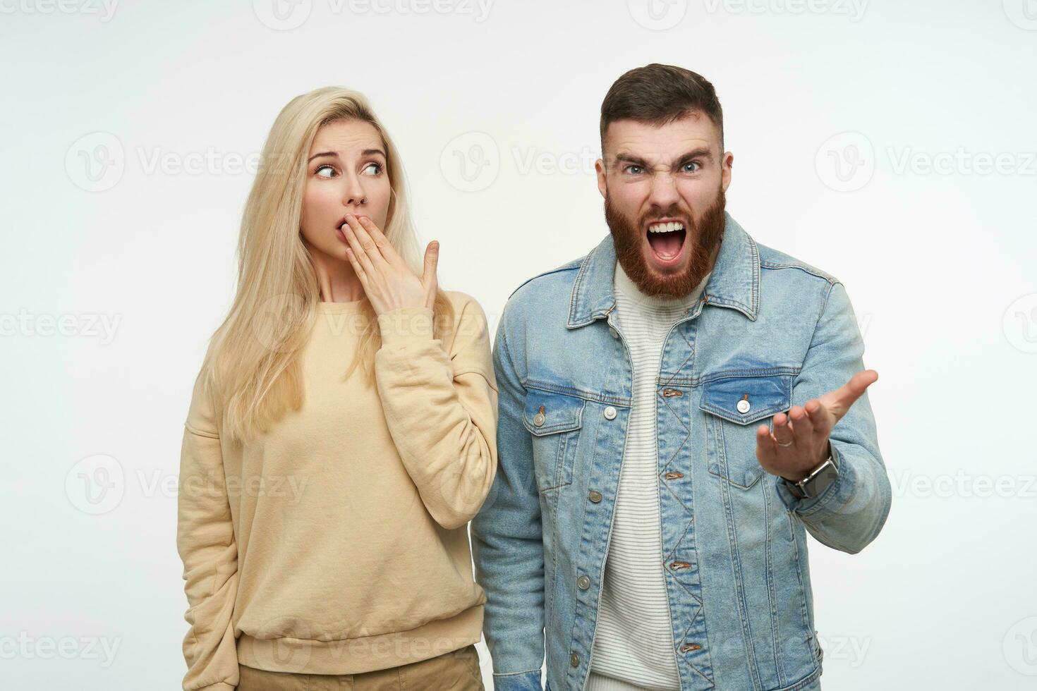 interior Disparo de joven asustado encantador rubia hembra mirando asustado en enojado barbado hermoso morena hombre gritando locamente, aislado terminado blanco antecedentes foto