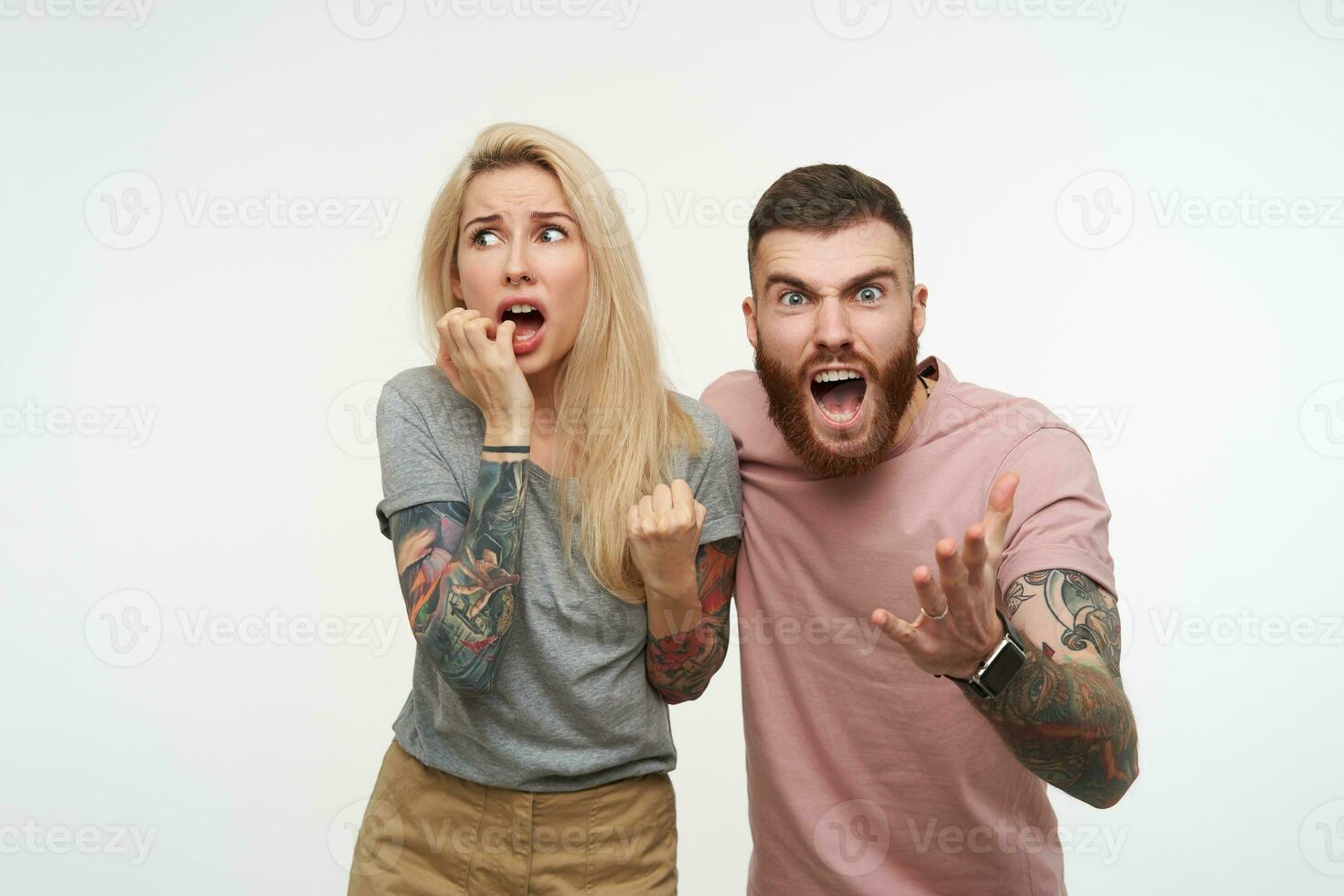 Excited young handsome brunette tattooed guy screaming excitedly and raising his hand while posing over white background with scared pretty blonde female in grey t-shirt photo