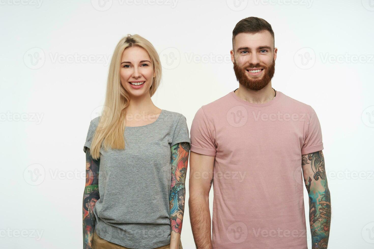 Studio photo of young lovely cheerful tattooed couple looking positively at camera with wide smiles and keeping their hands along bodies, posing over white background