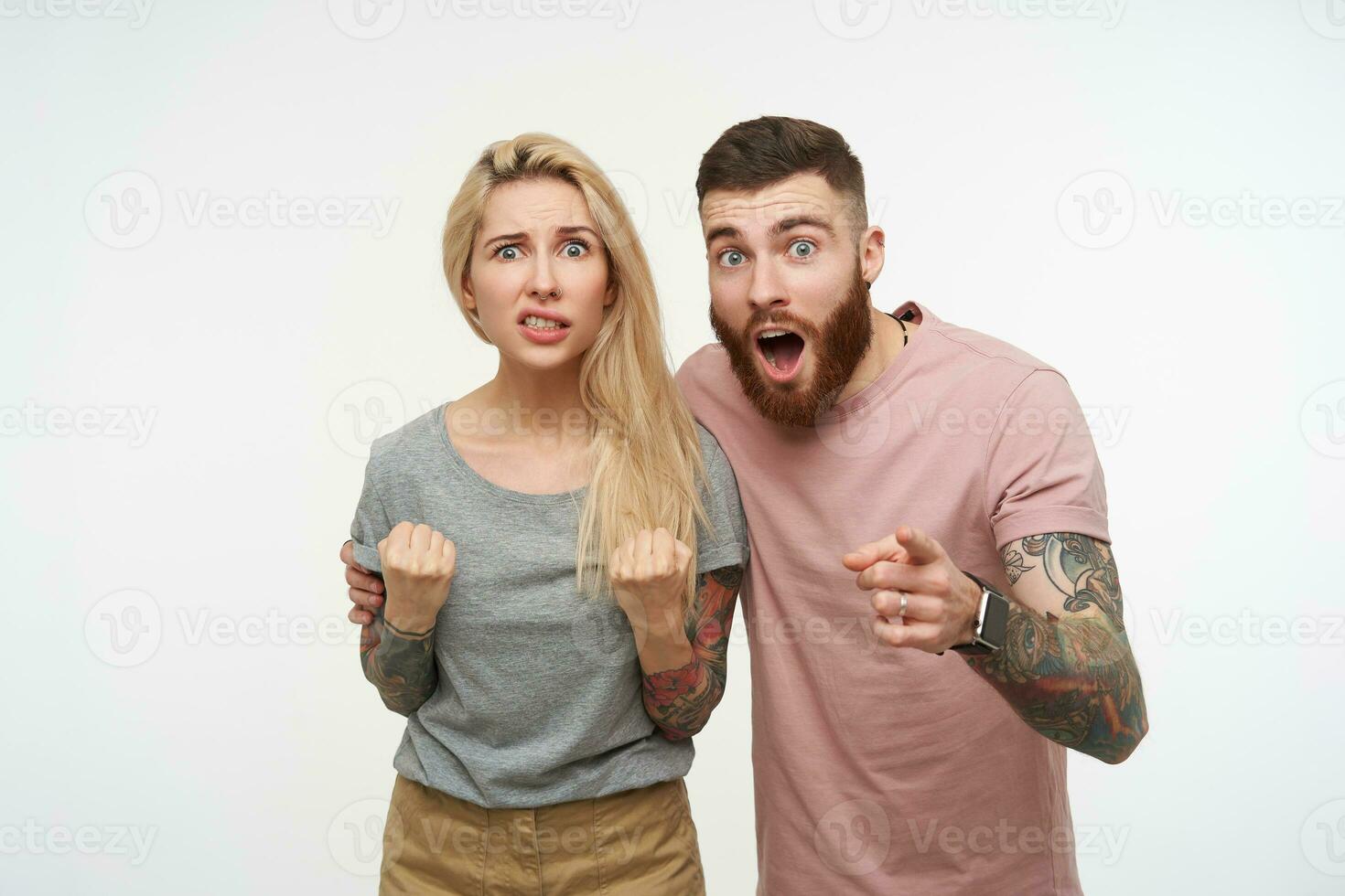 Confused young long haired blonde tattooed woman grimacing her face and keeping fists raised while posing over white background with her amazed brunette bearded boyfriend photo