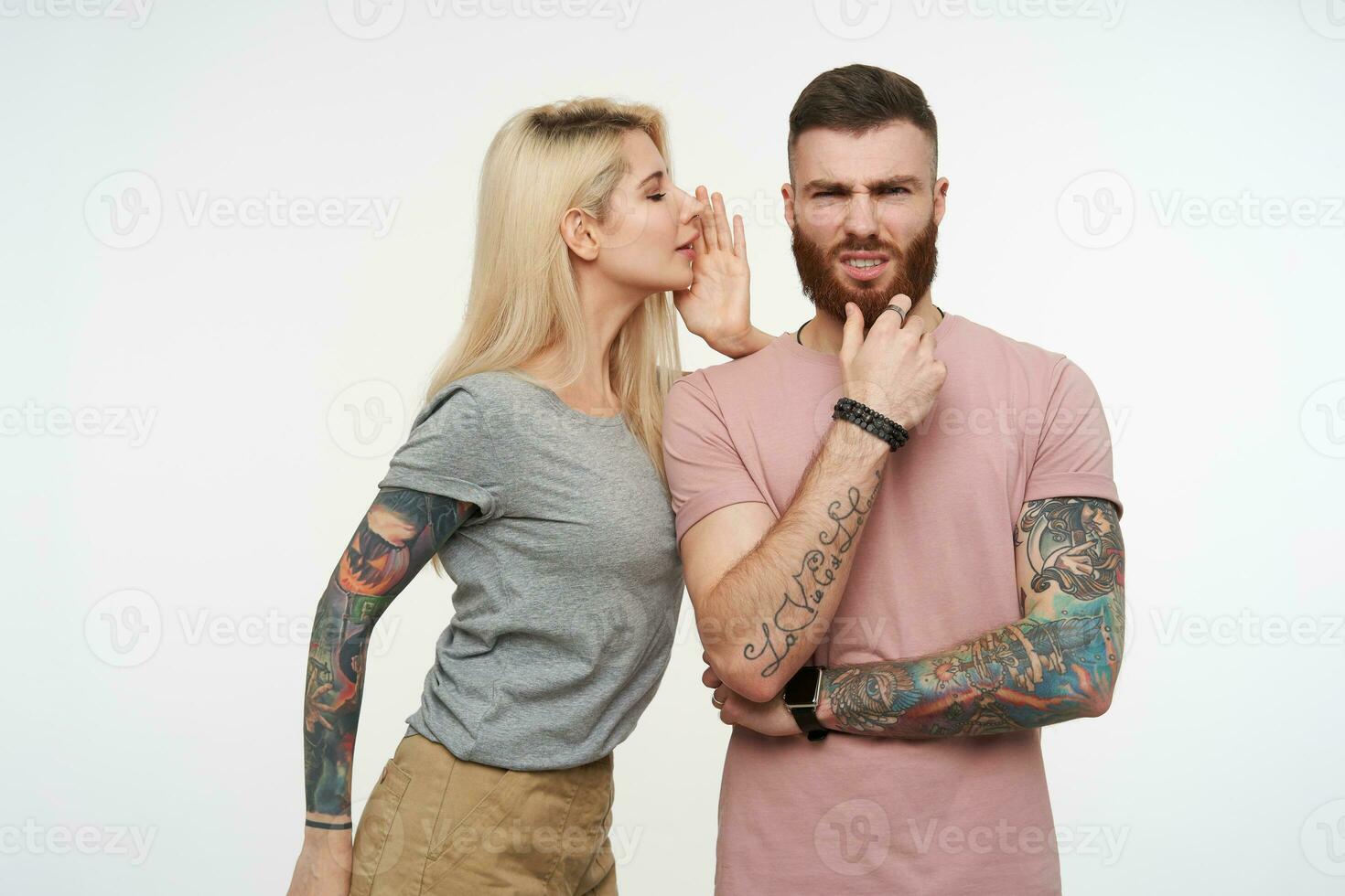 Studio photo of young attractive tattooed guy with beard frowning his face while looking pensively at camera and listening to his pretty long haired blonde girlfriend
