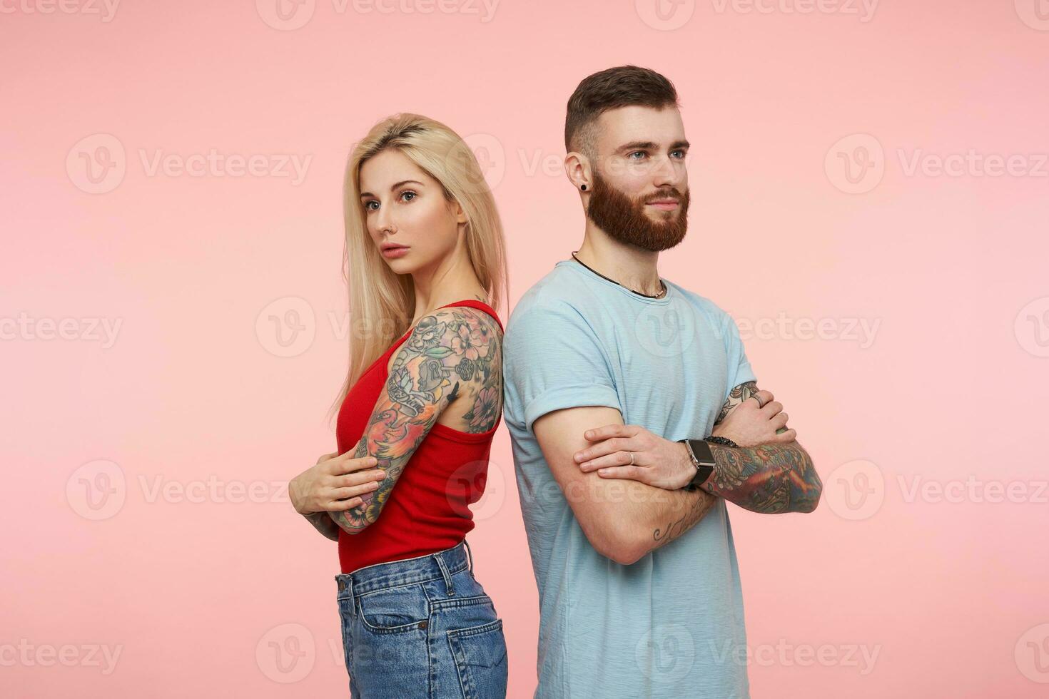 Pensive young pretty couple with tattooes crossing hands on their chest and looking thoughtfully with folded lips, standing over pink background in casual clothes photo