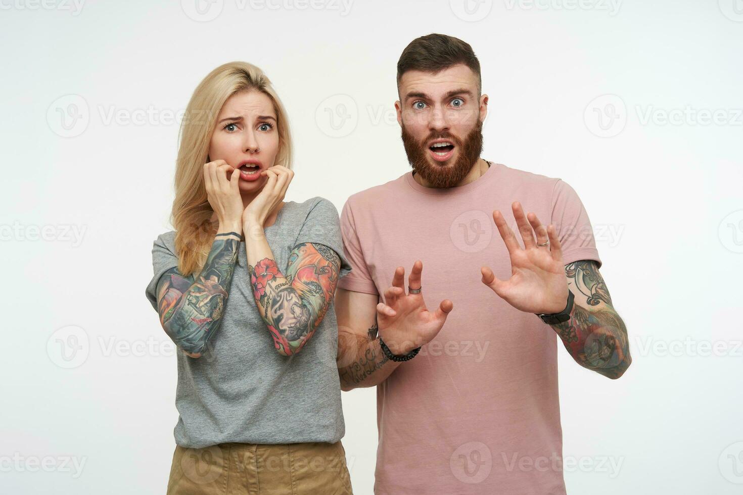 Frightened young attractive couple in casual basic t-shirts raising scaredly hands while looking wonderingly at camera, standing over white background photo