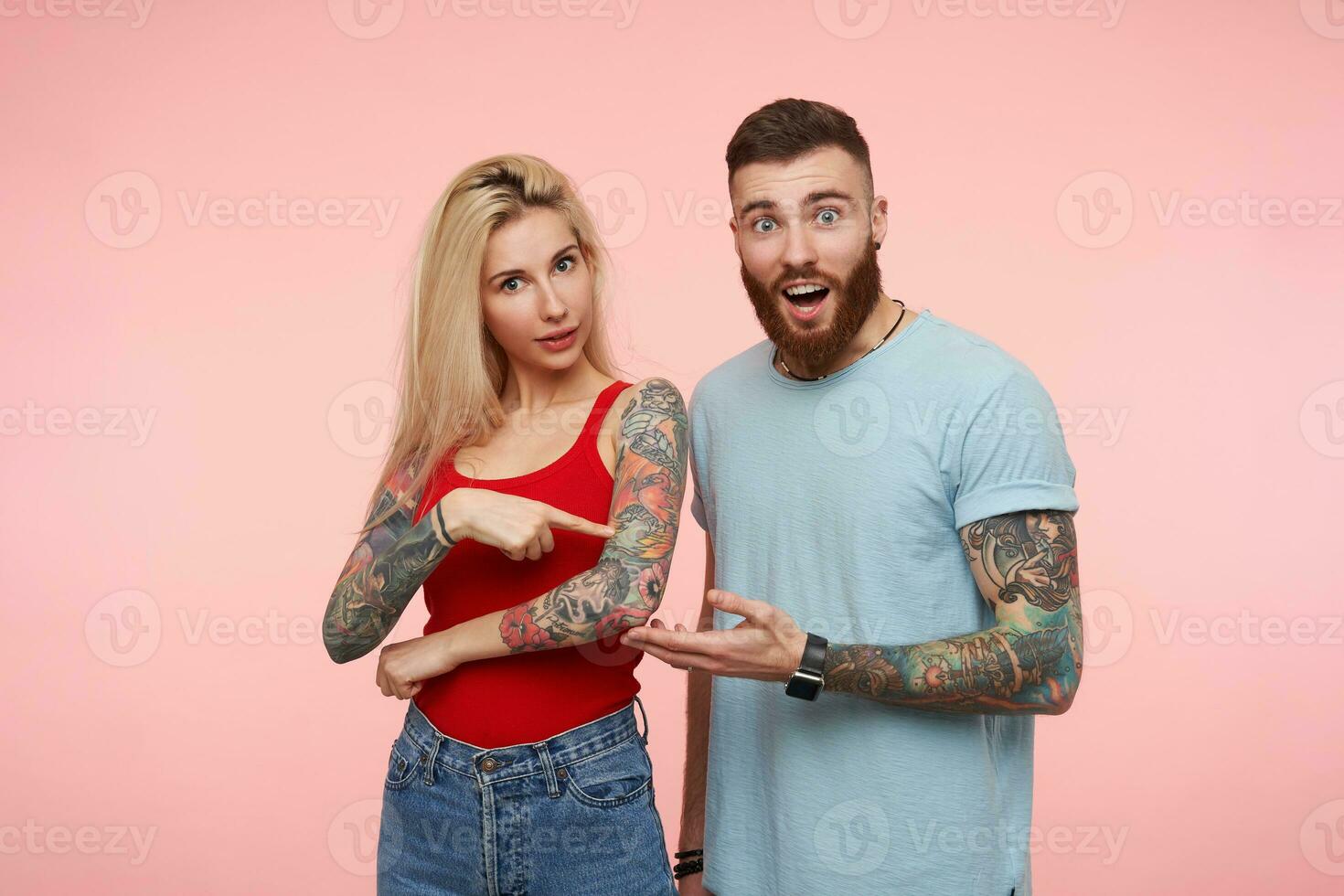Indoor photo of attractive couple dressed in casual clothes looking surprisedly at camera and keeping hands raised, standing against pink background