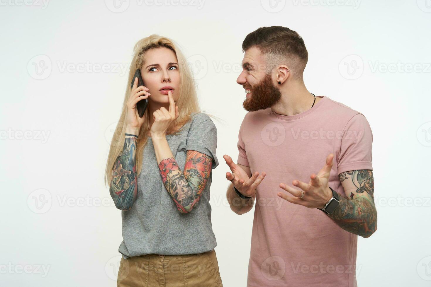 Studio photo of young long haired blonde lady looking pensively upwards while having phone talk, standing over white background with bewildered bearded tattooed male