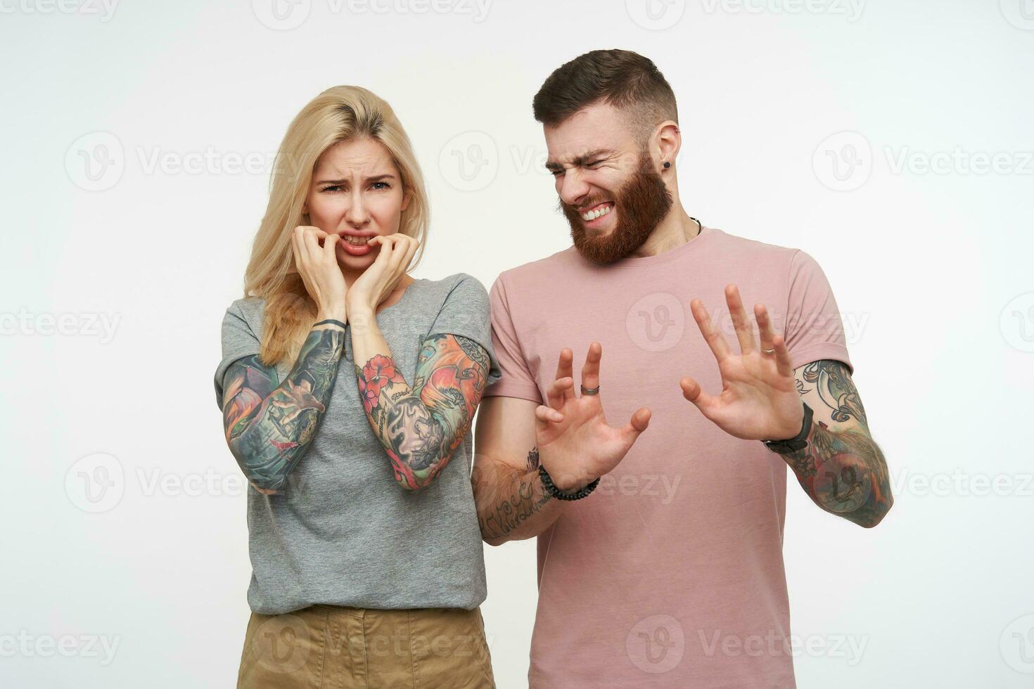 Indoor shot of young confused tattooed attractive couple keeping hands raised while grimacing their faces, being isolated over white background in casual wear photo