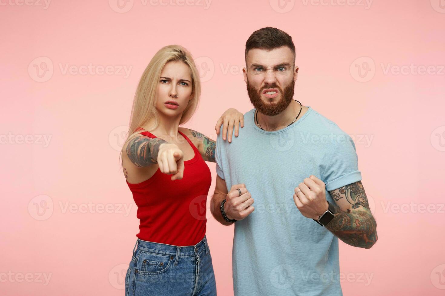 trastornado joven atractivo rubia mujer señalando con dedo índice adelante mientras posando terminado rosado antecedentes con su enojado pelo castaño tatuado hermoso amigo foto