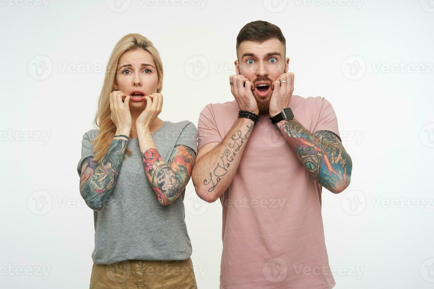 Shocked young pretty tattooed couple holding raised hands on their cheeks while looking amazedly at camera with wide eyes opened, isolated over white background photo