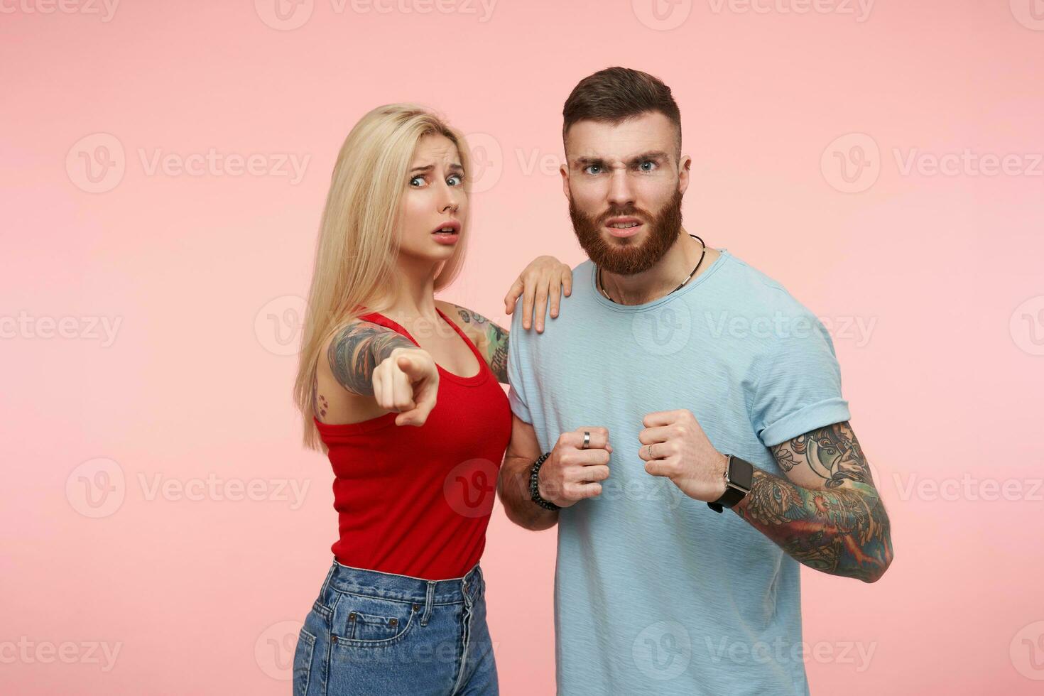 estudio foto de joven hermoso morena hombre levantamiento furiosamente su puño mientras yendo a proteger su trastornado bonito largo peludo tatuado rubia novia, aislado terminado rosado antecedentes