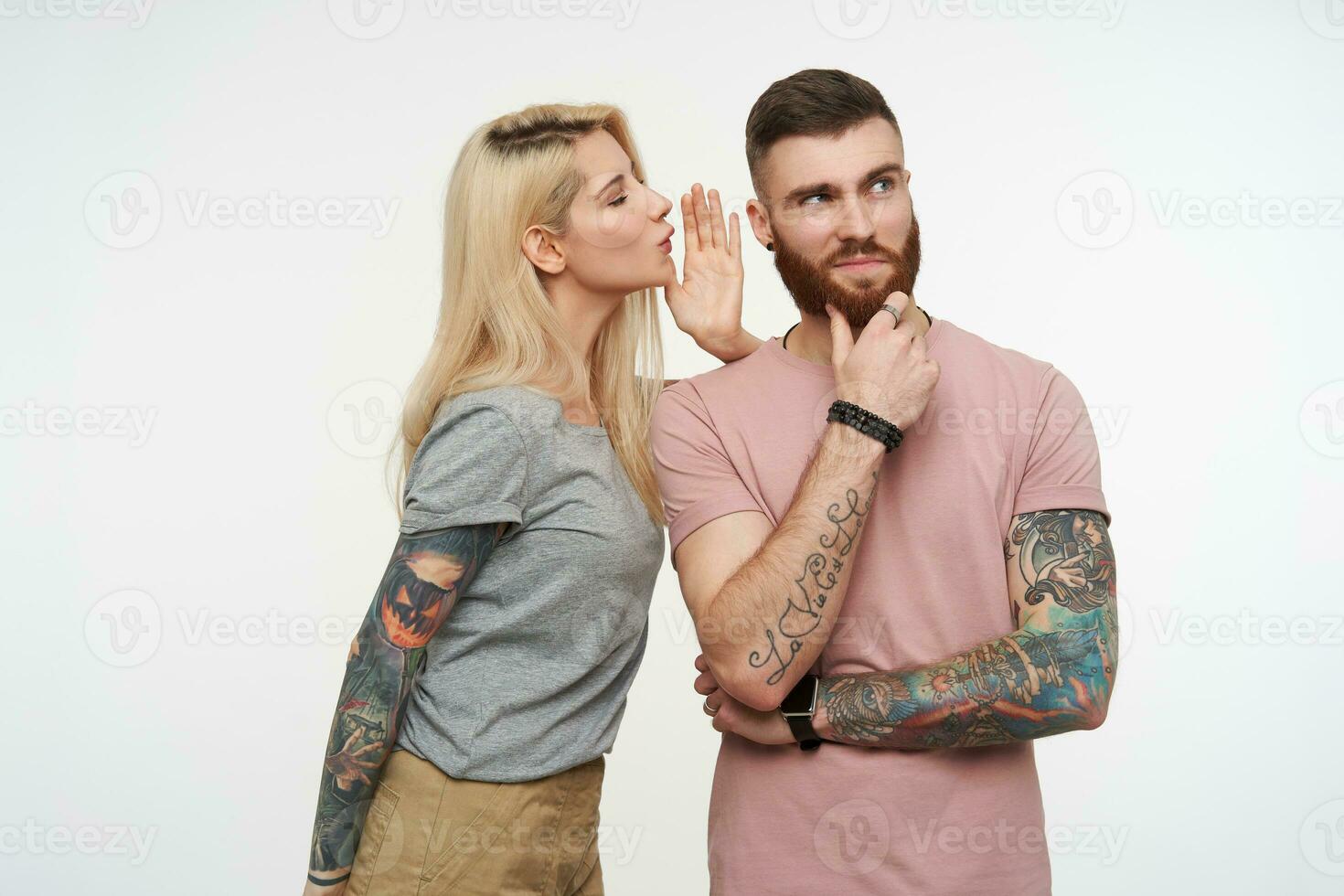 Positive young pretty bearded guy holding his chin with raised hand and looking dreamily upwards while pretty blonde tattooed female telling him something, isolated over white background photo