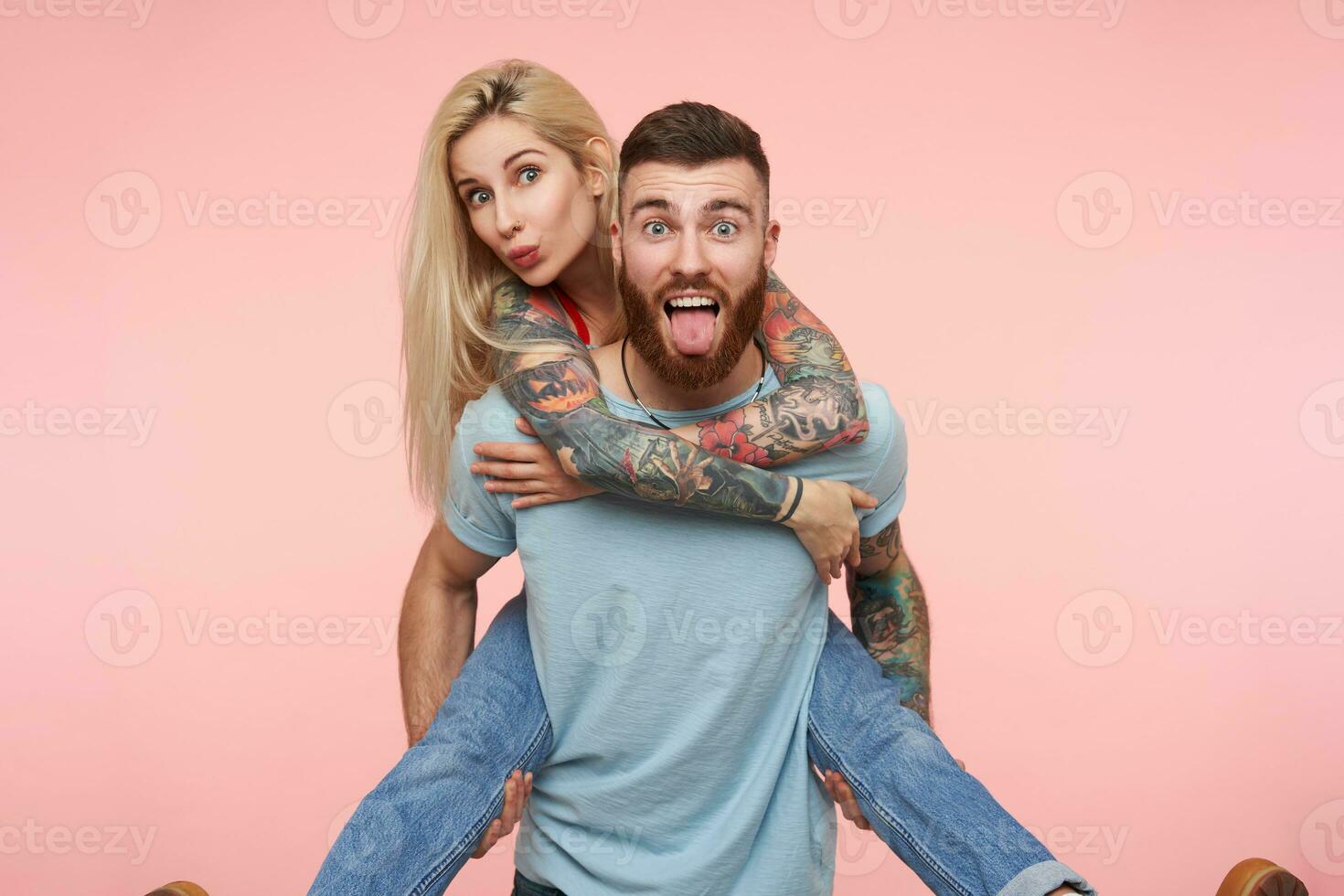 Horizontal shot of young excited pretty couple with tattooes looking joyfully at camera while fooling and making funny faces, standing over pink background photo