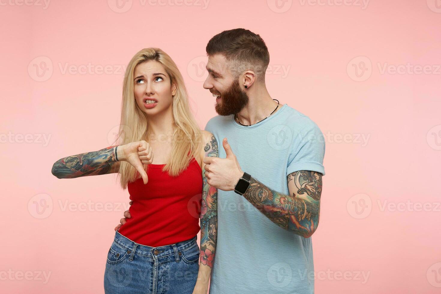 alegre joven atractivo barbado masculino en gris camiseta mirando alegremente a su rubia disgustado de pelo largo rubia Novia mientras en pie terminado rosado antecedentes foto