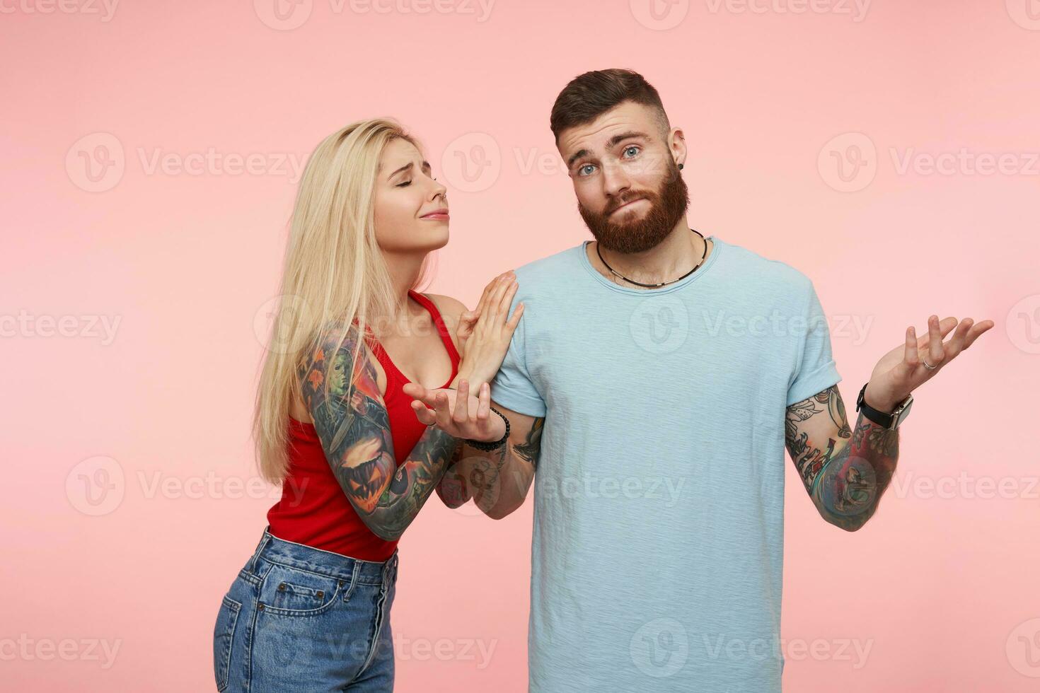 Puzzled young lovely bearded tattooed male shrugging with raised palms while his long haired pretty blonde girlfriend begging him for something, isolated over pink background photo