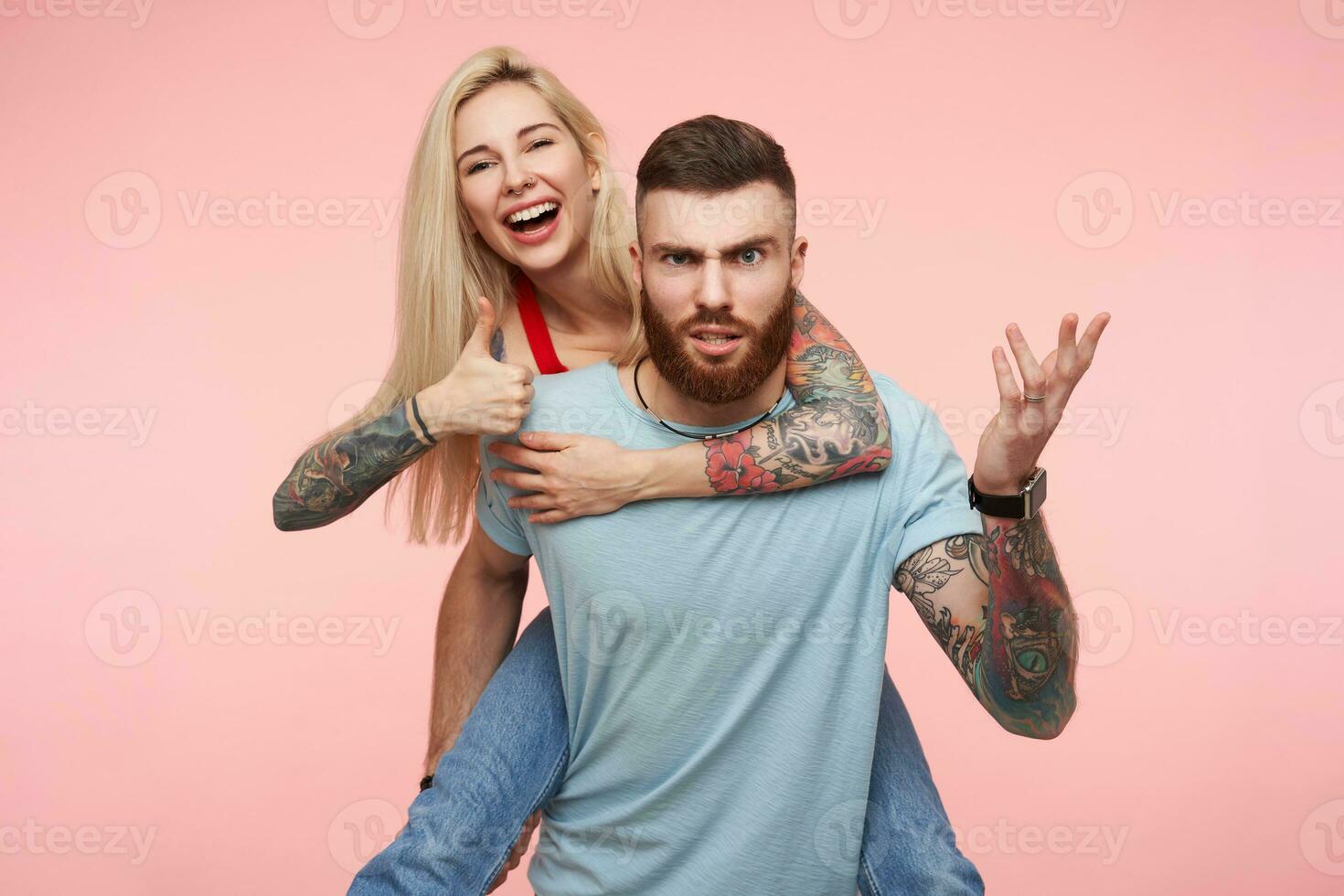 Severe young pretty bearded tattooed man frowning eyebrows while looking seriously at camera and keeping his cheerful attractive blonde girlfriend on his back, isolated over pink background photo