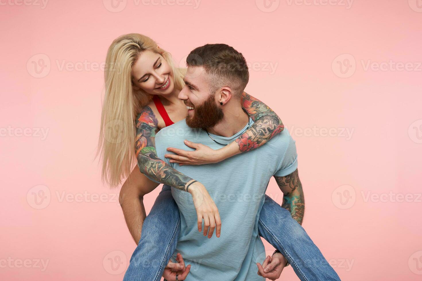 Studio photo of young cheerful attractive tattooed couple in casual clothes looking gladly on each other while having fun together over pink background