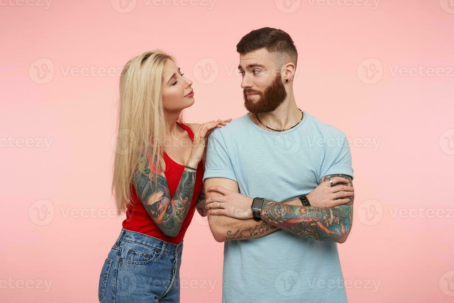 Positive young attractive tattooed male with beard crossing hands on chest and looking his long haired blonde pretty girlfriend, isolated over pink background photo