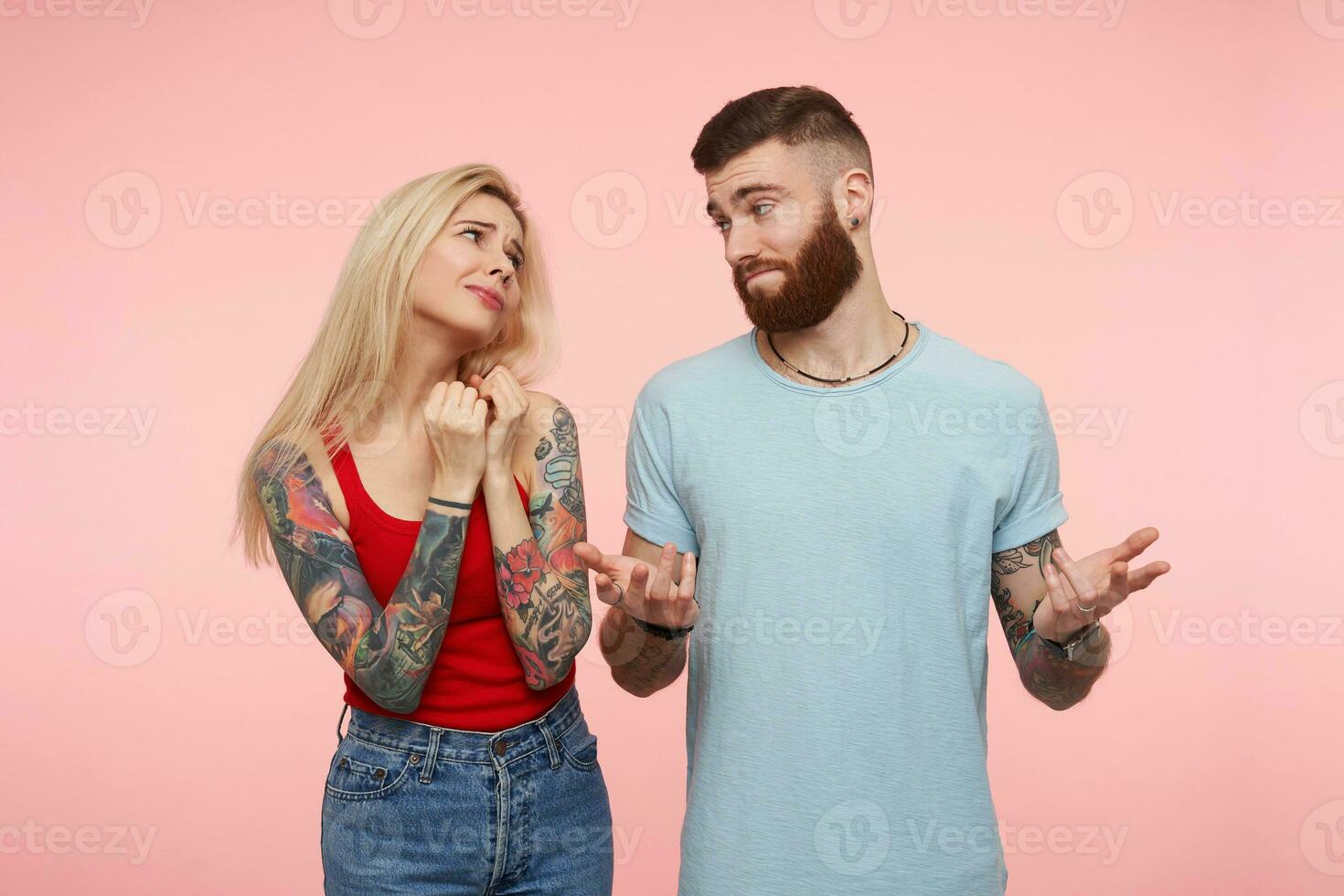 linda joven largo peludo rubia hembra con tatuajes plegable elevado manos mientras preguntando alguna cosa desde su bonito barbado tatuado amigo mientras siendo aislado terminado rosado antecedentes foto