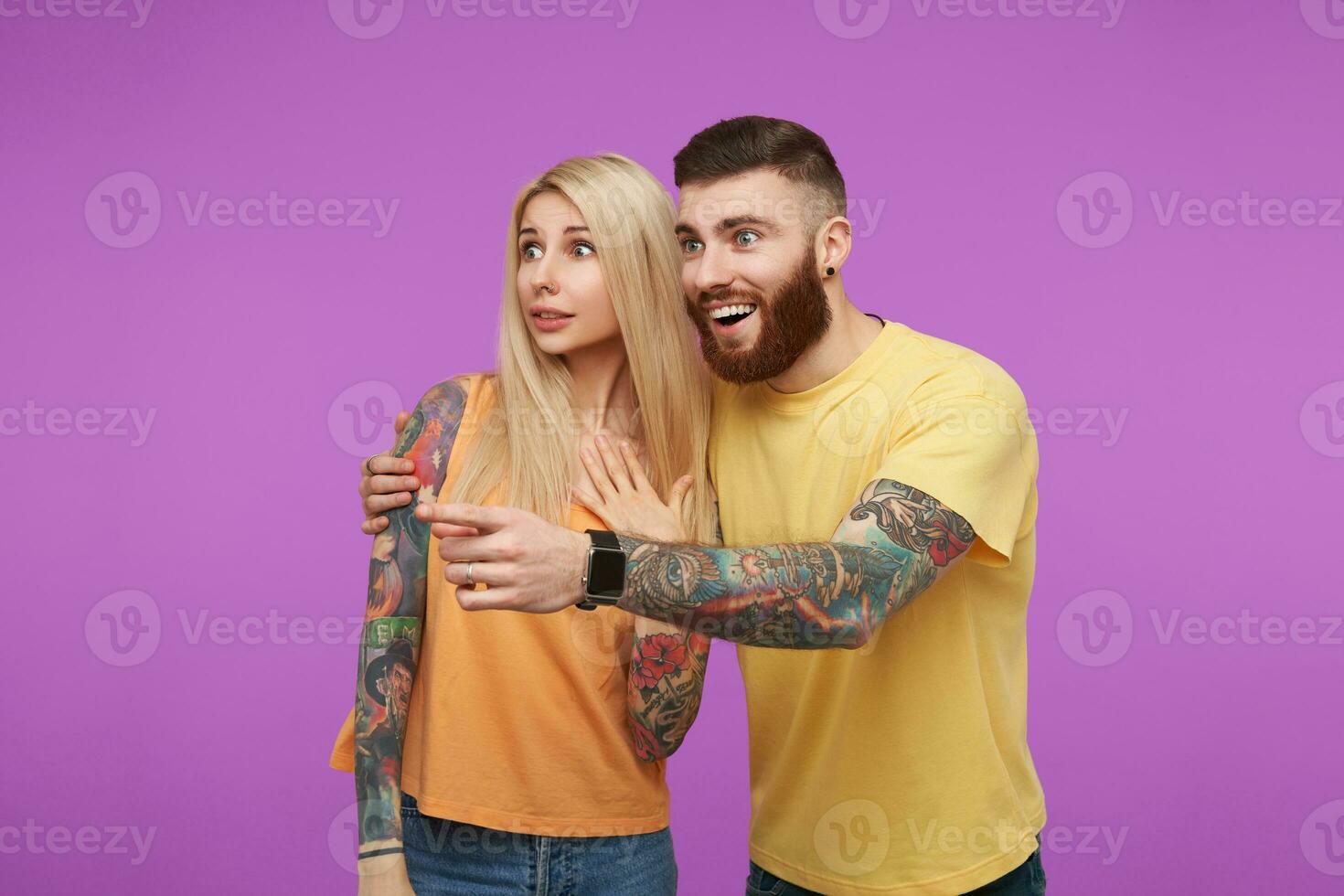 Surprised young lovely people in orange casual clothes rounding wonderingly their blue eyes while looking aside, standing against purple background photo