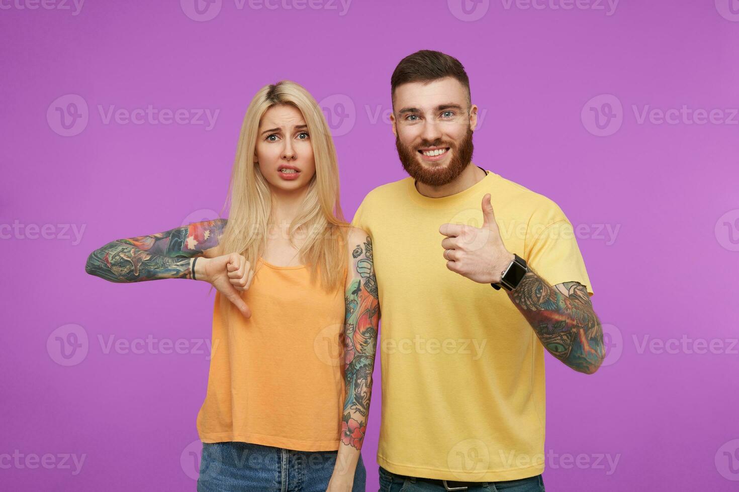 Joyful young tattooed bearded male smiling cheerfully while thumbing up, posing over purple background with long haired blonde dissatisfied woman showing dislike sign photo