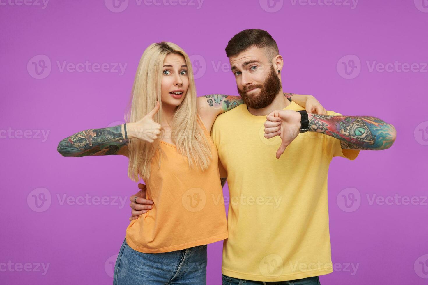 estudio foto de joven de ojos azules tatuado bonito Pareja demostración me gusta y disgusto mientras mirando a cámara y abrazando cada otro, aislado terminado púrpura antecedentes