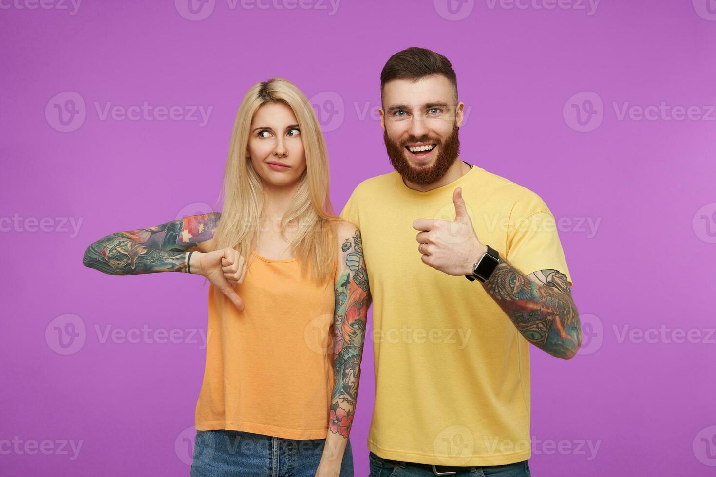 Studio photo of attractive pair of young people with tattooes gesturing with raised hands while demonstrating different moods, standing over purple background